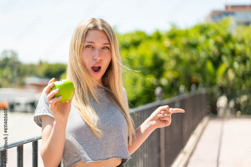 Young blonde woman with an apple at outdoors surprised and pointing side Young blonde woman with an apple at outdoors surprised and pointing side