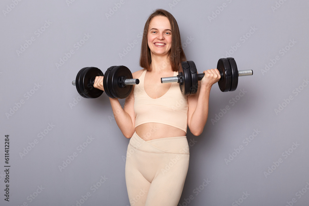 Smiling attractive charming positive woman wearing top holding barbells in hands isolated on gray background having workout for arms enjoying sport trainings. Smiling attractive charming positive woman wearing top holding barbells in hands isolated on gray background having workout for arms enjoying sport trainings.