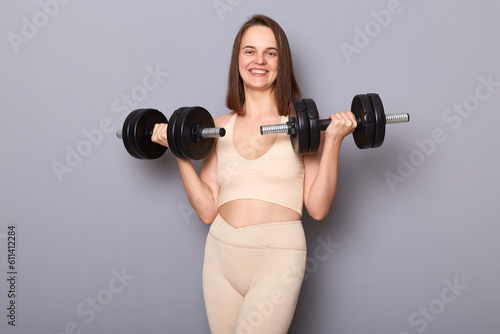 Smiling attractive charming positive woman wearing top holding barbells in hands isolated on gray background having workout for arms enjoying sport trainings.