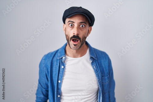 Hispanic man with beard standing over isolated background in shock face, looking skeptical and sarcastic, surprised with open mouth