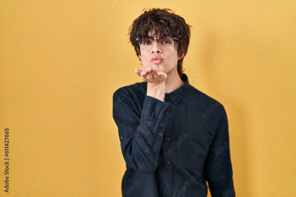 Young man wearing glasses over yellow background looking at the camera blowing a kiss with hand on air being lovely and sexy. love expression.