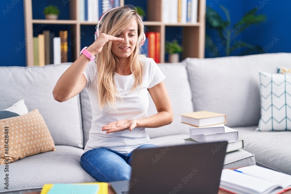 Young blonde woman studying using computer laptop at home gesturing with hands showing big and large size sign, measure symbol. smiling looking at the camera. measuring concept. Young blonde woman studying using computer laptop at home gesturing with hands showing big and large size sign, measure symbol. smiling looking at the camera. measuring concept.