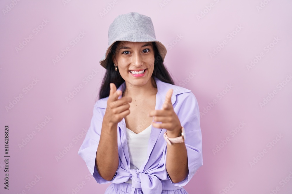 Young hispanic woman standing over pink background wearing hat pointing fingers to camera with happy and funny face. good energy and vibes. Young hispanic woman standing over pink background wearing hat pointing fingers to camera with happy and funny face. good energy and vibes.