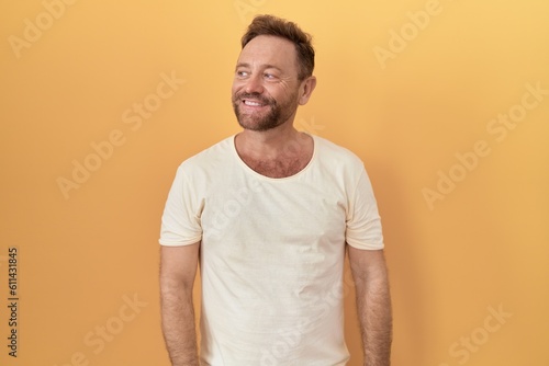 Middle age man with beard standing over yellow background smiling looking to the side and staring away thinking.