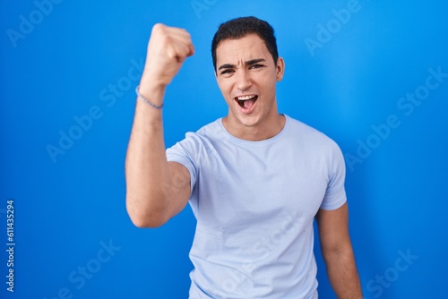 Young hispanic man standing over blue background angry and mad raising fist frustrated and furious while shouting with anger. rage and aggressive concept.