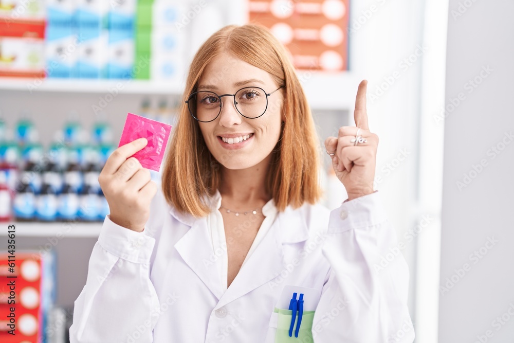 Young redhead woman working at pharmacy drugstore holding condom surprised with an idea or question pointing finger with happy face, number one