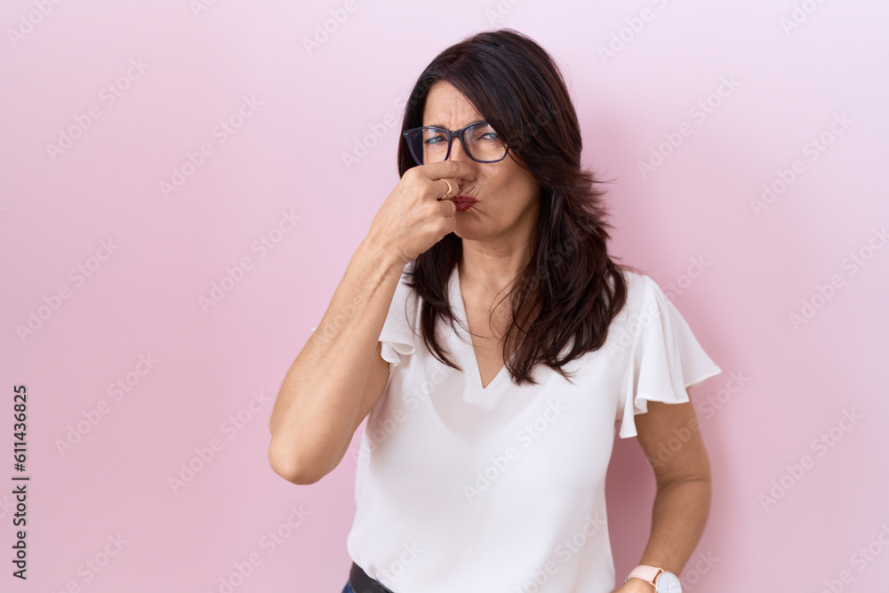 Middle age hispanic woman wearing casual white t shirt and glasses smelling something stinky and disgusting, intolerable smell, holding breath with fingers on nose. bad smell Middle age hispanic woman wearing casual white t shirt and glasses smelling something stinky and disgusting, intolerable smell, holding breath with fingers on nose. bad smell