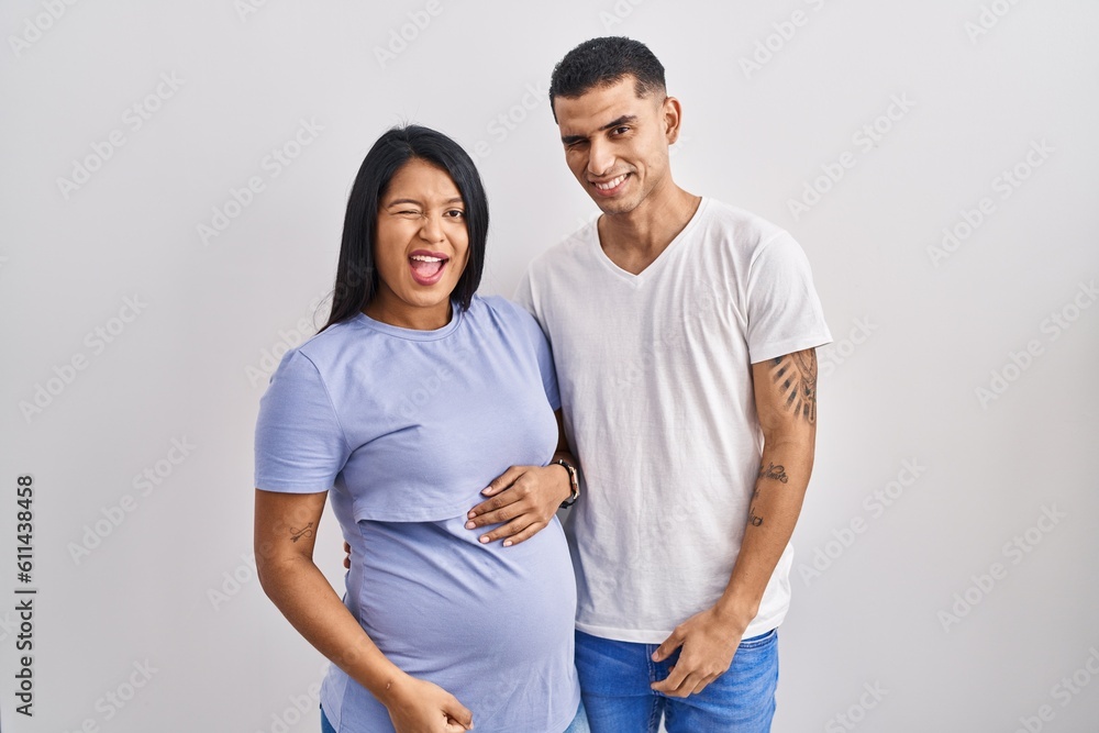 Young hispanic couple expecting a baby standing over background winking looking at the camera with sexy expression, cheerful and happy face. Young hispanic couple expecting a baby standing over background winking looking at the camera with sexy expression, cheerful and happy face.
