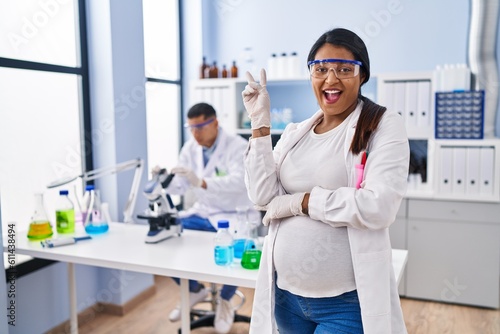 Young hispanic woman expecting a baby working at scientist laboratory smiling with happy face winking at the camera doing victory sign. number two.