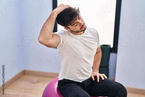 Young hispanic man sitting on fit ball stretching at sport center