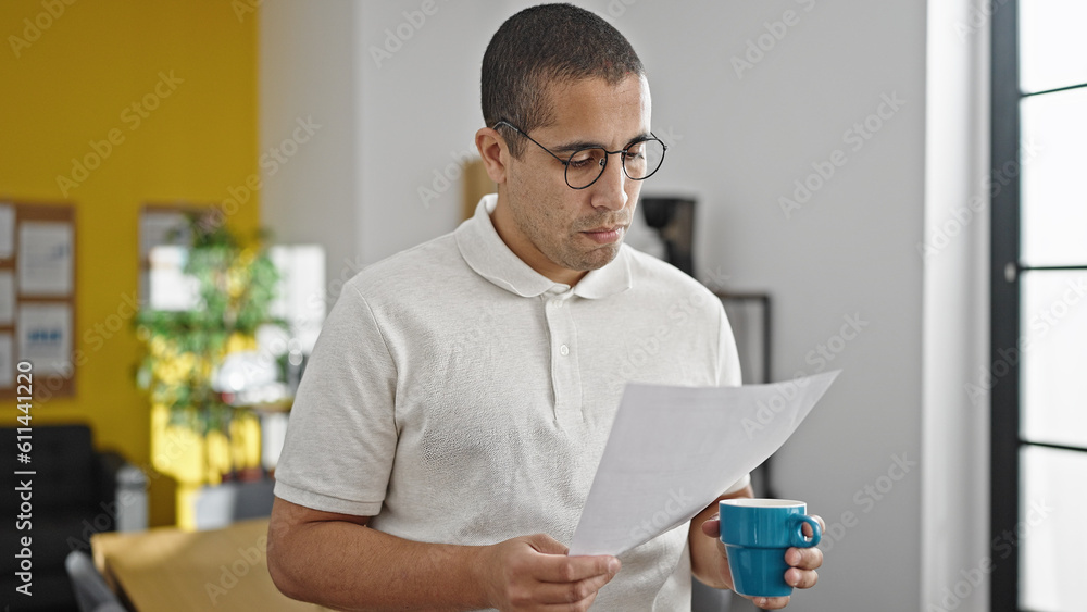 Fototapeta premium Young hispanic man business worker drinking coffee reading document at office