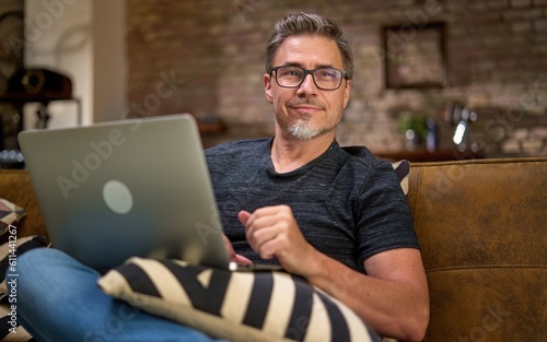 Older man working online with laptop computer at home sitting on couch in living room. Home office, browsing internet. Portrait of happy, mature age, middle age, mid adult man in 50s, smiling.