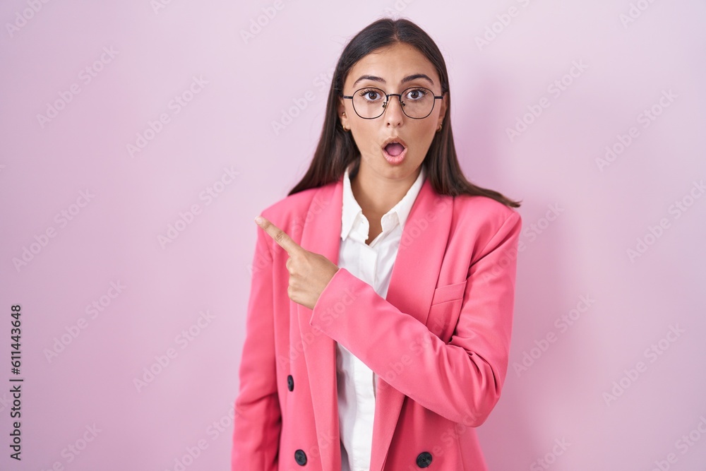 Young hispanic woman wearing business clothes and glasses surprised pointing with finger to the side, open mouth amazed expression. Young hispanic woman wearing business clothes and glasses surprised pointing with finger to the side, open mouth amazed expression.