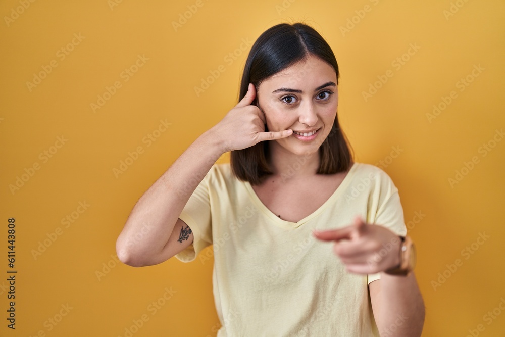 Hispanic girl wearing casual t shirt over yellow background smiling doing talking on the telephone gesture and pointing to you. call me. Hispanic girl wearing casual t shirt over yellow background smiling doing talking on the telephone gesture and pointing to you. call me.