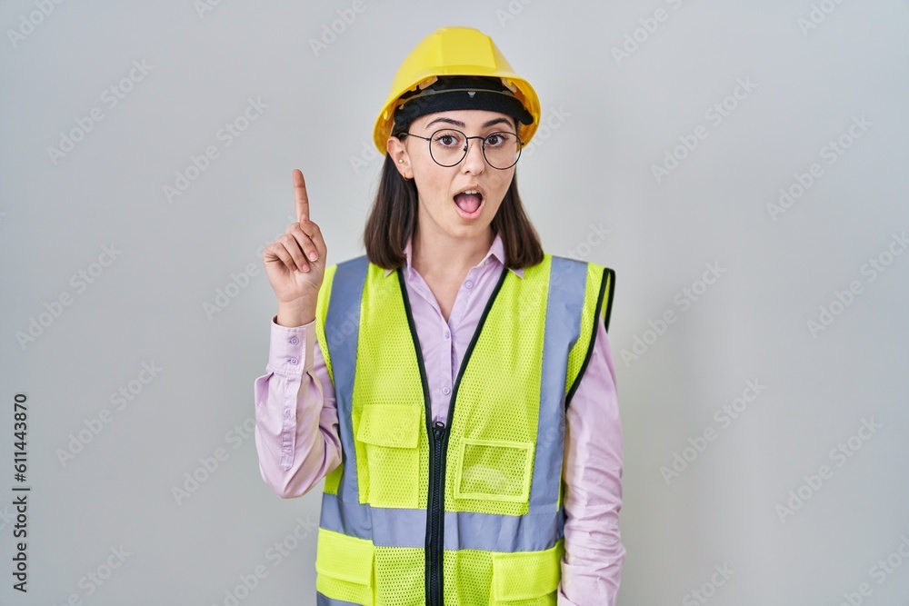 Hispanic girl wearing builder uniform and hardhat pointing finger up with successful idea. exited and happy. number one.