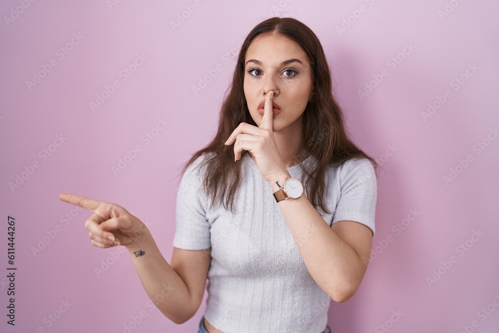 Young hispanic girl standing over pink background asking to be quiet with finger on lips pointing with hand to the side. silence and secret concept. Young hispanic girl standing over pink background asking to be quiet with finger on lips pointing with hand to the side. silence and secret concept.