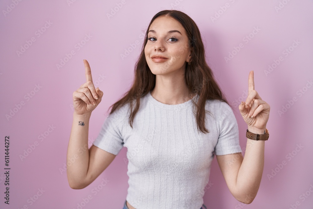 Young hispanic girl standing over pink background smiling amazed and surprised and pointing up with fingers and raised arms. Young hispanic girl standing over pink background smiling amazed and surprised and pointing up with fingers and raised arms.