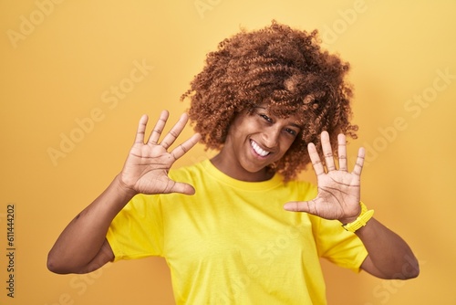Young hispanic woman with curly hair standing over yellow background showing and pointing up with fingers number ten while smiling confident and happy.