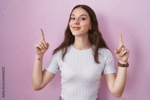 Young hispanic girl standing over pink background smiling amazed and surprised and pointing up with fingers and raised arms.