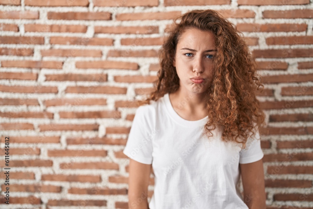 Young caucasian woman standing over bricks wall background puffing cheeks with funny face. mouth inflated with air, crazy expression.
