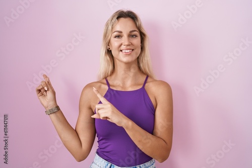 Young blonde woman standing over pink background smiling and looking at the camera pointing with two hands and fingers to the side.