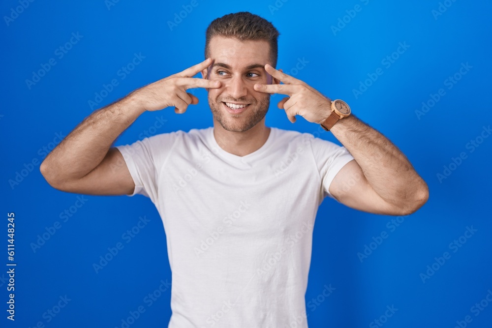 Naklejka premium Young caucasian man standing over blue background doing peace symbol with fingers over face, smiling cheerful showing victory