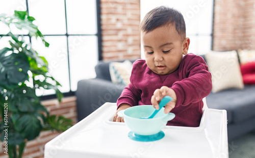 Adorable hispanic boy sitting on highchair holding spoon at home