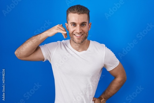 Young caucasian man standing over blue background smiling doing phone gesture with hand and fingers like talking on the telephone. communicating concepts.