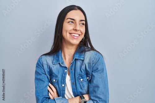 Hispanic woman standing over blue background looking away to side with smile on face, natural expression. laughing confident.