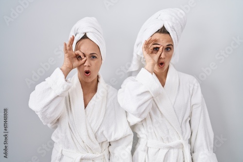 Middle age woman and daughter wearing white bathrobe and towel doing ok gesture shocked with surprised face, eye looking through fingers. unbelieving expression.