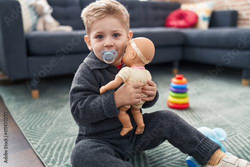 Fotografiet Adorable caucasian boy playing with baby doll sitting on floor at home