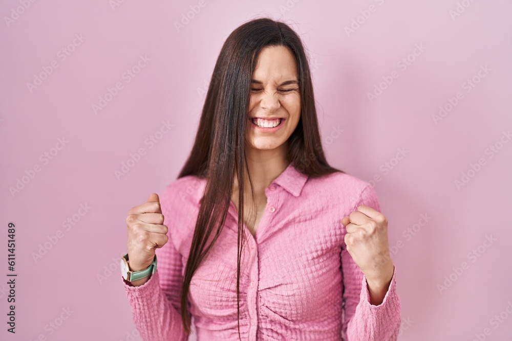 Young hispanic woman standing over pink background very happy and excited doing winner gesture with arms raised, smiling and screaming for success. celebration concept. Young hispanic woman standing over pink background very happy and excited doing winner gesture with arms raised, smiling and screaming for success. celebration concept.