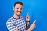 Young hispanic man standing over blue background smiling and looking at the camera pointing with two hands and fingers to the side.