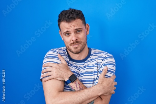 Young hispanic man standing over blue background shaking and freezing for winter cold with sad and shock expression on face