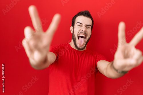 Young hispanic man wearing casual red t shirt smiling with tongue out showing fingers of both hands doing victory sign. number two. © Krakenimages.com