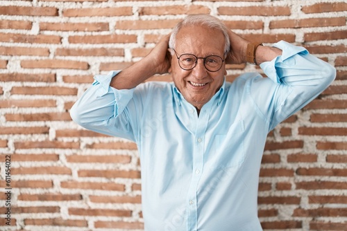 Senior man with grey hair standing over bricks wall relaxing and stretching, arms and hands behind head and neck smiling happy