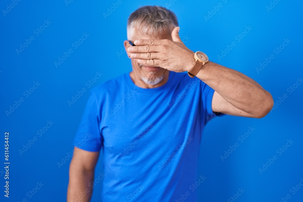 Hispanic man with grey hair standing over blue background smiling and laughing with hand on face covering eyes for surprise. blind concept. Hispanic man with grey hair standing over blue background smiling and laughing with hand on face covering eyes for surprise. blind concept.