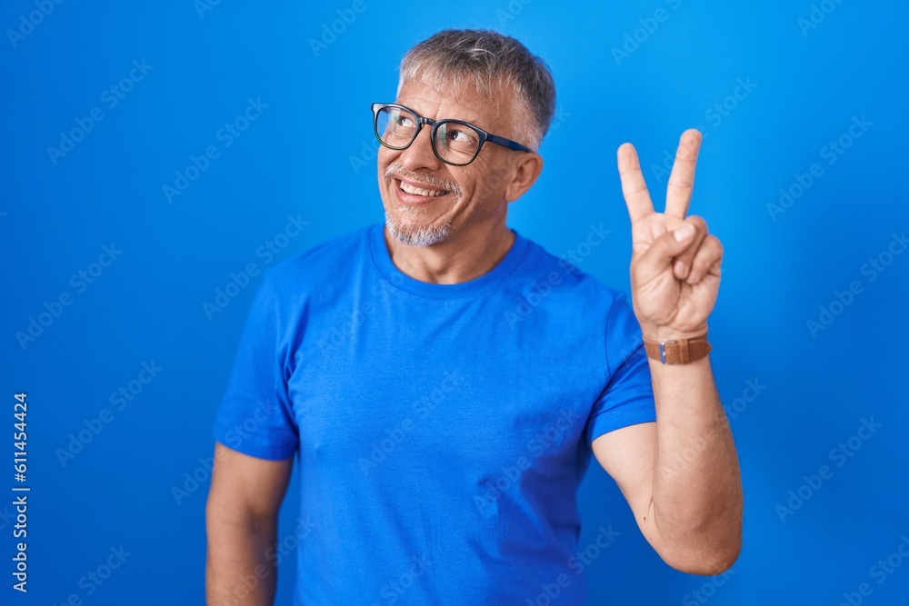 Hispanic man with grey hair standing over blue background smiling looking to the camera showing fingers doing victory sign. number two. Hispanic man with grey hair standing over blue background smiling looking to the camera showing fingers doing victory sign. number two.