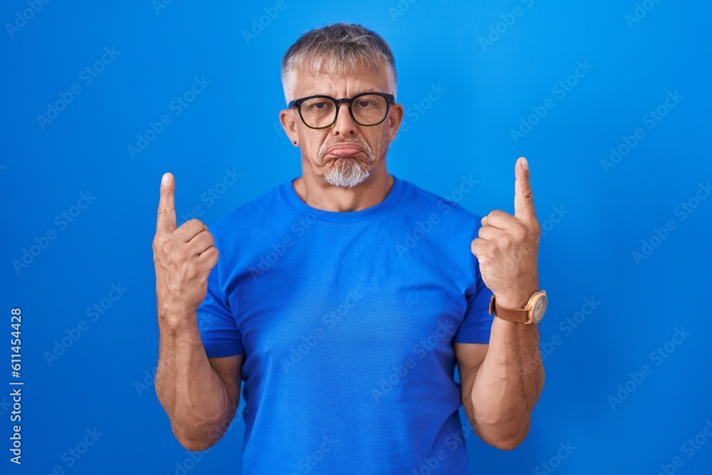 Hispanic man with grey hair standing over blue background pointing up looking sad and upset, indicating direction with fingers, unhappy and depressed. Hispanic man with grey hair standing over blue background pointing up looking sad and upset, indicating direction with fingers, unhappy and depressed.