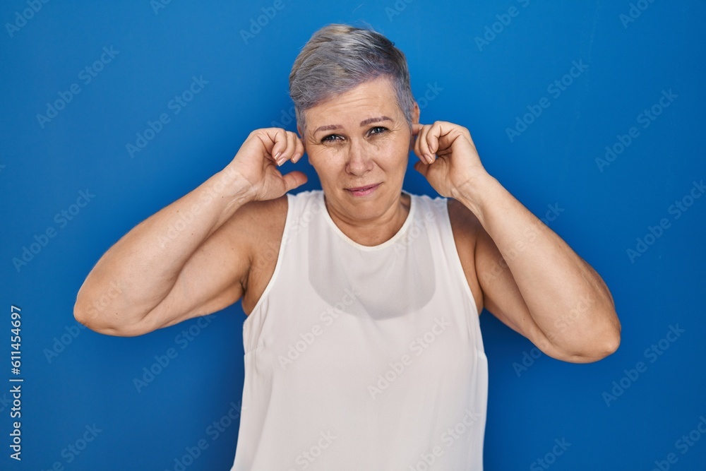 Middle age caucasian woman standing over blue background covering ears with fingers with annoyed expression for the noise of loud music. deaf concept. Middle age caucasian woman standing over blue background covering ears with fingers with annoyed expression for the noise of loud music. deaf concept.