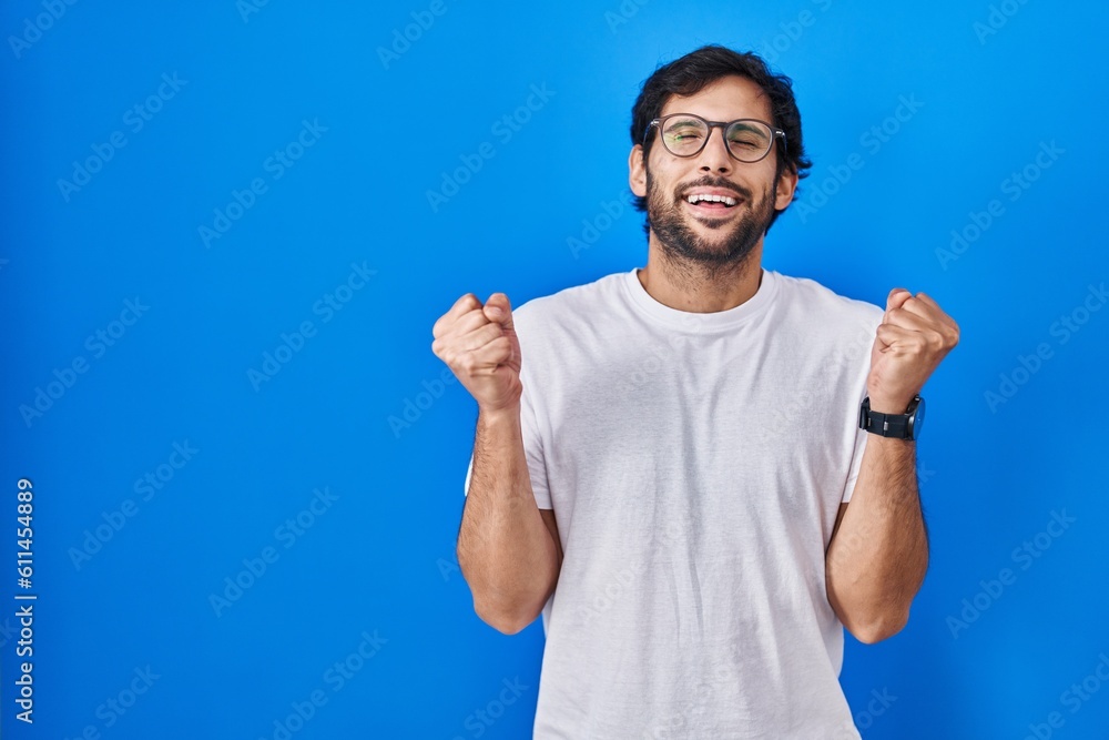 Handsome latin man standing over blue background excited for success with arms raised and eyes closed celebrating victory smiling. winner concept. Handsome latin man standing over blue background excited for success with arms raised and eyes closed celebrating victory smiling. winner concept.