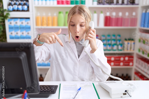 Young caucasian woman working at pharmacy drugstore speaking on the telephone pointing down with fingers showing advertisement, surprised face and open mouth