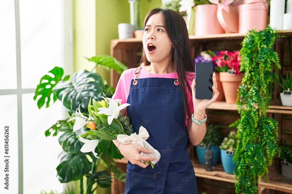 Young chinese woman working at florist shop showing smartphone screen angry and mad screaming frustrated and furious, shouting with anger looking up.