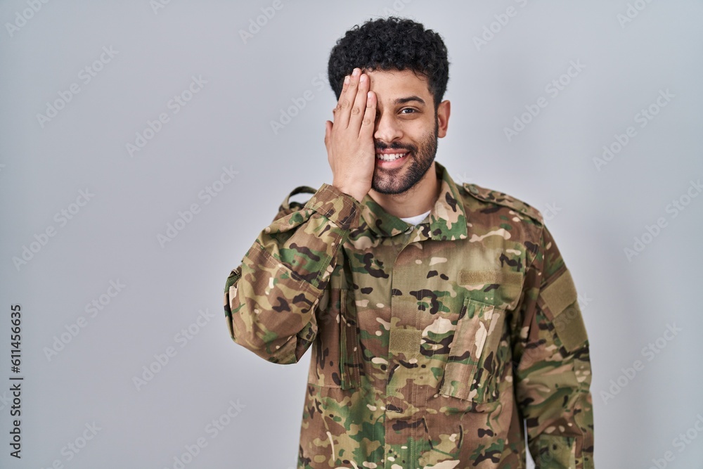 Arab man wearing camouflage army uniform covering one eye with hand, confident smile on face and surprise emotion.