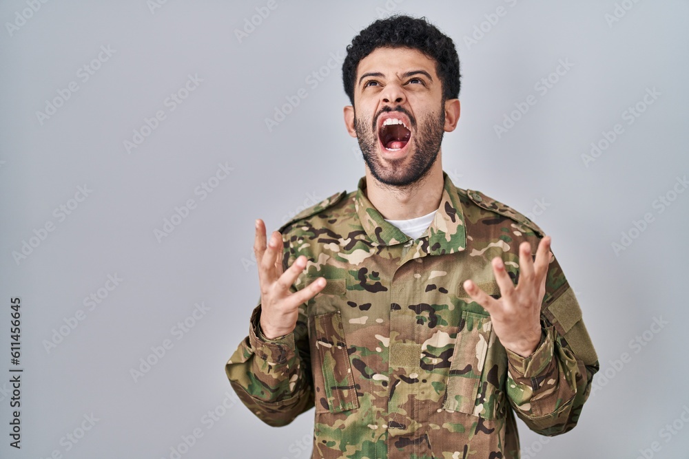 Arab man wearing camouflage army uniform crazy and mad shouting and yelling with aggressive expression and arms raised. frustration concept. Arab man wearing camouflage army uniform crazy and mad shouting and yelling with aggressive expression and arms raised. frustration concept.