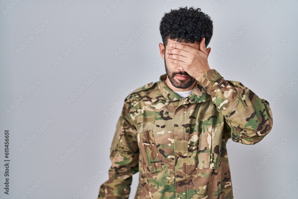 Arab man wearing camouflage army uniform covering eyes with hand, looking serious and sad. sightless, hiding and rejection concept