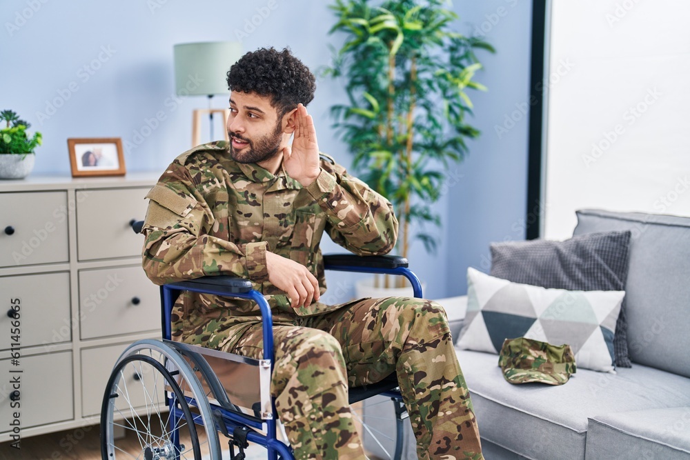 Arab man wearing camouflage army uniform sitting on wheelchair smiling with hand over ear listening an hearing to rumor or gossip. deafness concept.