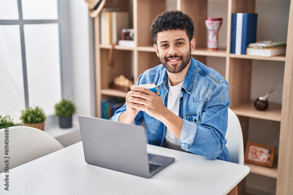 Young arab man using laptop drinking coffe sitting on table at home Young arab man using laptop drinking coffe sitting on table at home