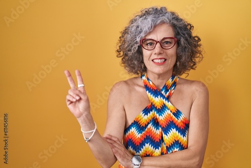 Middle age woman with grey hair standing over yellow background smiling with happy face winking at the camera doing victory sign with fingers. number two.