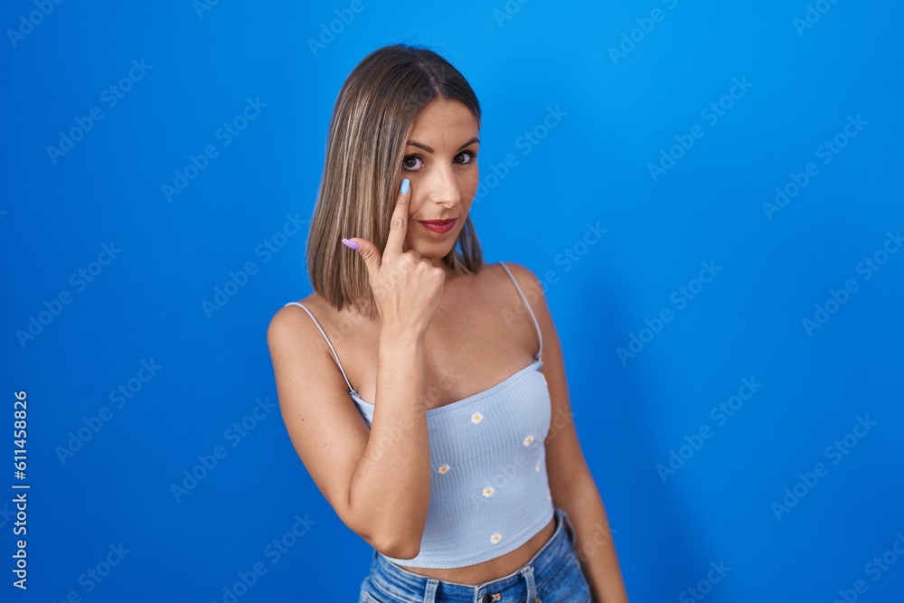 Fototapeta premium Young woman standing over blue background pointing to the eye watching you gesture, suspicious expression